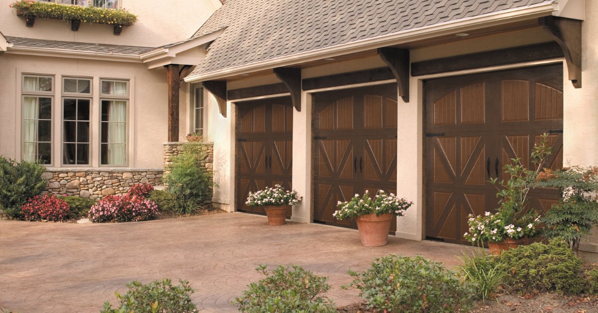 A tan house with two brown garage doors, flower planters, bushes, and a paved driveway near the front windows.