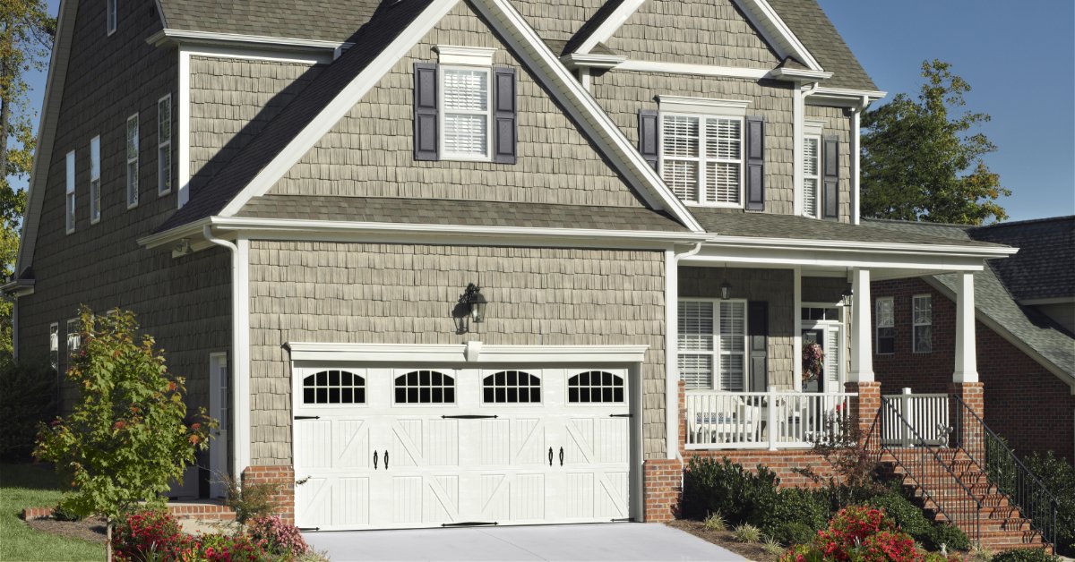 A gray house with a white garage door, front porch, windows, landscaped shrubs, and a clear blue sky.